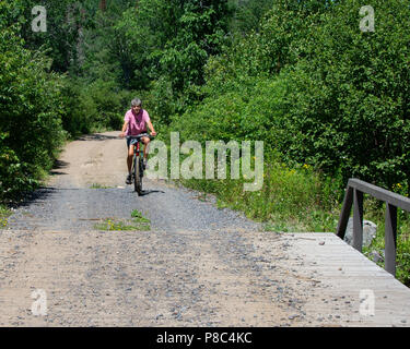 Senior woman riding a bicycle on a dirt and gravel road in the Adirondack Mountains, NY USA Stock Photo