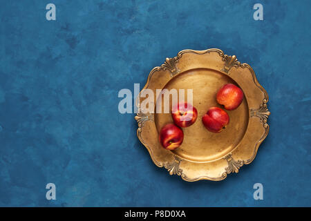Flat lay of fresh nectarines fruits on rustic golden serving plate on blue background. Top view with copy space. Stock Photo