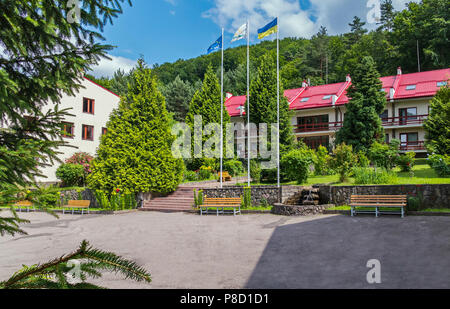 Comfortable wooden benches, a decorative stone fountain and tall fir trees at the entrance to the hotel building with a red roof . For your design Stock Photo