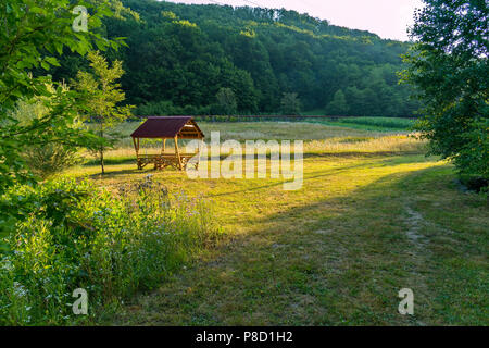 Arbor for rest with a table standing on a green lawn next to the country gardens with growing crops on them . For your design Stock Photo