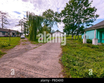 Paths among low-rise buildings in the countryside with lawn-growing willows bending their branches to the ground and a car standing side by side. . Fo Stock Photo