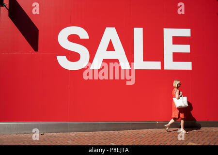People out shoppin walk past a large scale sale sign in white lettering on a red background outside H&M, a major high street clothing retail shop in Birmingham, United Kingdom. Its time for the summer sales, and most shops are advertising big reductions in prices. Bargains are avaiable and the shopping streets are busy. Stock Photo