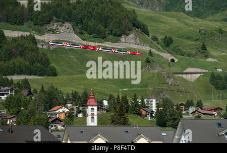 Andermatt in the Canton of Uri, Switzerland, showing Swiss Moutain Trains-Glacier Express- climbing up towards Oberalp Pass and Swiss flags. June 2018 Stock Photo