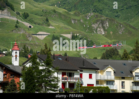 Andermatt in the Canton of Uri, Switzerland, showing Swiss Moutain Trains-Glacier Express- climbing up towards Oberalp Pass and Swiss flags. June 2018 Stock Photo