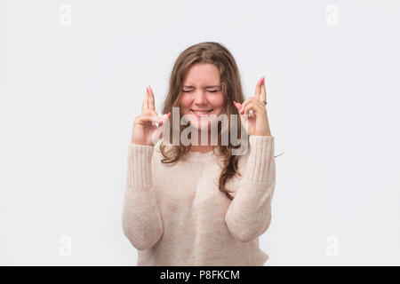 Studio portrait of attractive european woman in red shirt having excited, superstitious and naive look, keeping fingers crossed, hoping to win lottery Stock Photo