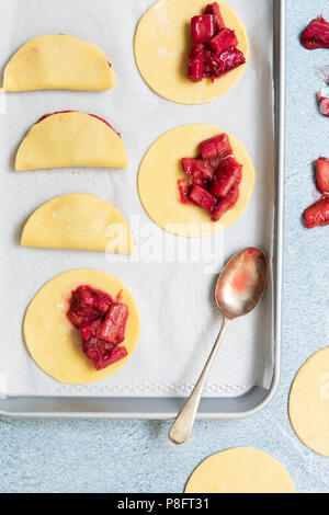 Preparation of roasted rhubarb turnovers, spoonfuls of filling placed on rounds of shortcrust pastry with three completed. Stock Photo