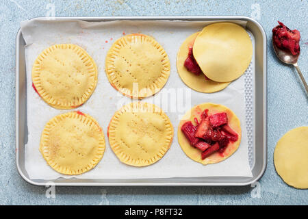 Uncooked roasted rhubarb turnovers on a baking tray, with a spoonful of filling and one uncovered. Stock Photo