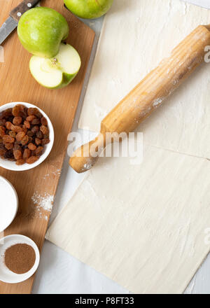 Ingredients for apple studel, including sugar, cinnamon, apples and sultanas on a wooden cutting board with a rolling pin and two pastry sheets. Stock Photo