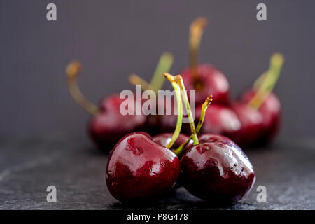 Closeup of three juicy ripe dark red cherries in foreground on rustic slate surface with out of focus bunch against black background Stock Photo