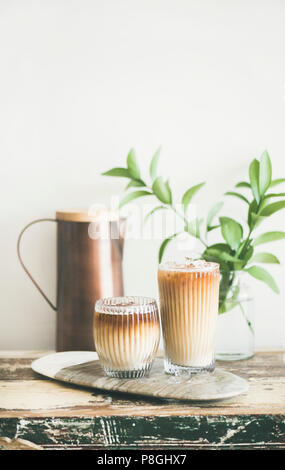 Iced coffee in tall glasses with milk on board, white wall, green plant branches and jug at background, copy space. Summer refreshing beverage ice cof Stock Photo