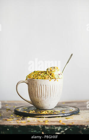 Homemade pistachio ice cream in ceramic mug with green pistachio nuts on rustic wooden table, vertical composition, copy space. Summer refreshing ice Stock Photo