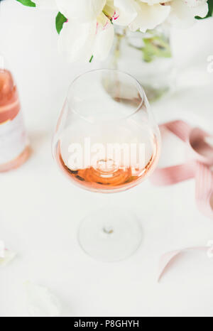Rose wine in glass, pink decorative ribbon, peony flowers over white background, selective focus, close-up. Summer celebration, wedding greeting card, Stock Photo