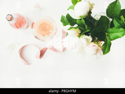 Flat-lay of rose wine in glass and bottle, pink decorative ribbon and bouquet of peony flowers over white table background, top view, copy space. Summ Stock Photo