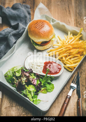 Classic burger dinner. Beef meat homemade burger with French fries, salad and sauces on white tray over rustic wooden table background Stock Photo