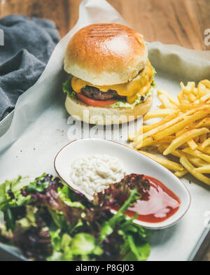 Classic burger dinner. Beef meat homemade burger with French fries, salad and sauces on white tray over rustic wooden table background, selective focu Stock Photo