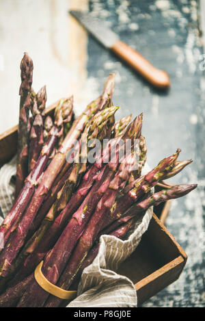 Seasonal harvest produce. Fresh raw uncooked purple asparagus over wooden tray background, selective focus, vertical composition. Local market food co Stock Photo