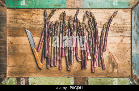 Seasonal harvest produce. Flat-lay of fresh raw uncooked purple asparagus in row over wooden tray background, top view. Local market food concept Stock Photo