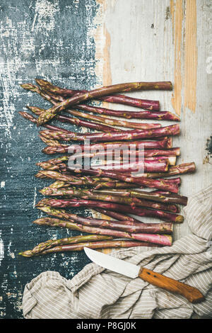Seasonal harvest produce. Flat-lay of fresh raw uncooked purple asparagus in row over rustic wooden background, top view, copy space, vertical composi Stock Photo