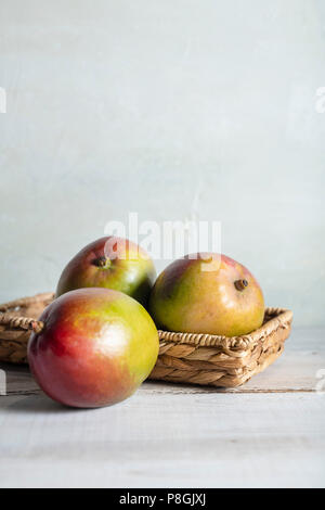Three ripe mangoes in a wicker basket on a whitewash wood table with bright, natural light.