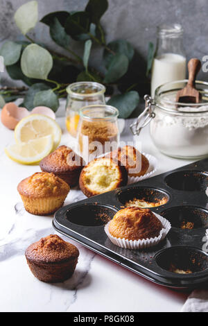 Fresh baked homemade lemon cakes muffins standing in baking dish with eucalyptus branch and ingredients above over white marble kitchen table. Stock Photo