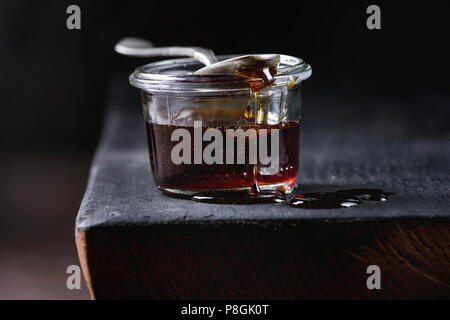 Homemade liquid transparent brown sugar caramel in glass jar standing on black wooden board with spoon. Close up Stock Photo