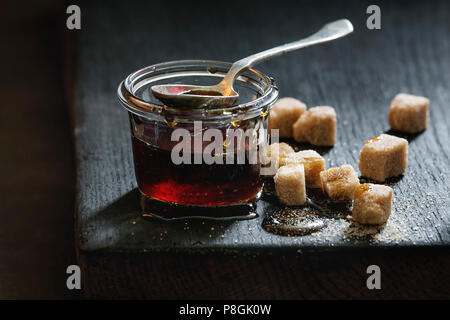 Homemade liquid transparent brown sugar caramel in glass jar standing on black wooden board with spoon and can sugar cubes. Close up. Day light Stock Photo