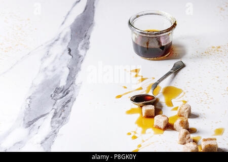 Homemade liquid transparent brown sugar caramel in glass jar with spoon and cane sugar cubes over white marble background. Copy space. Stock Photo