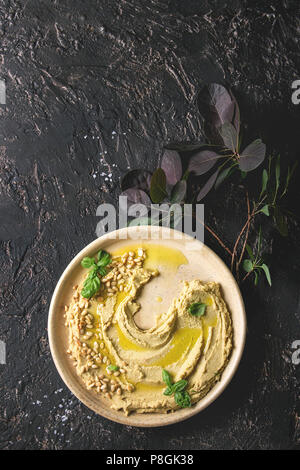 Homemade traditional spread hummus with pine nuts, olive oil, basil served on ceramic plate with green branch over dark texture background. Flat lay, Stock Photo