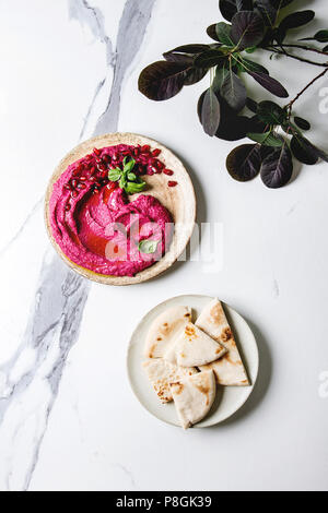 Homemade spread beetroot hummus with pomegranate seeds, olive oil, basil served on ceramic plate with pita bread and green branch over white marble ba Stock Photo