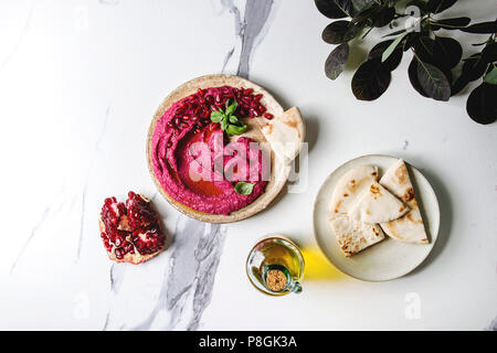 Homemade spread beetroot hummus with pomegranate seeds, olive oil, basil served on ceramic plate with pita bread and green branch over white marble ba Stock Photo