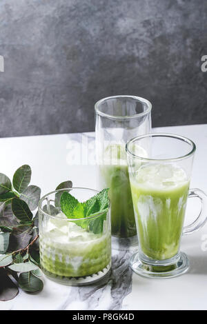 Matcha green tea iced latte or cocktail in three different glasses with ice cubes, matcha powder and jug of milk on white marble table, decorated by g Stock Photo
