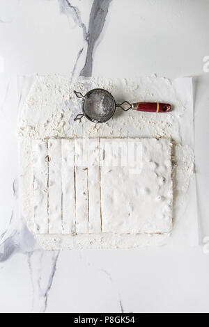 Homemade honey nuts nougat turron sliced on crumpled paper with vintage sieve and sugar powder over white marble background. Flat lay, space. Stock Photo