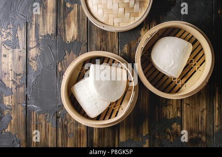 Empty gua bao steamed buns in opened bamboo steamer over dark wooden plank background. Flat lay, space. Asian fast food.
