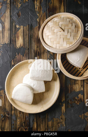 Empty gua bao steamed buns in ceramic plate and opened bamboo steamer over dark wooden plank background. Flat lay, space. Asian fast food.
