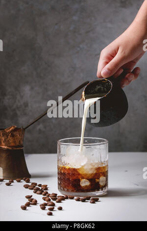 Iced coffee cocktail or frappe with ice cubes and cream, pouring from jug, served in glass with vintage jezva and coffee beans around on white marble Stock Photo