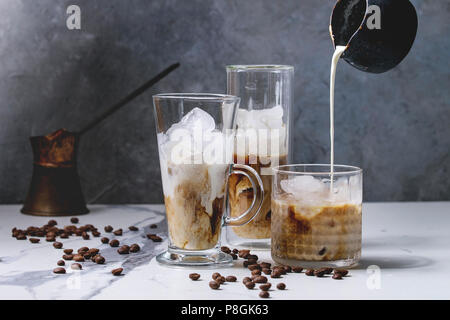 Iced coffee cocktail or frappe with ice cubes and pouring cream in three different glasses with vintage jezva and coffee beans around on white marble Stock Photo