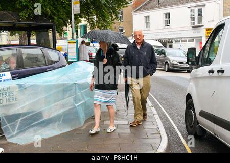 Axminster, Devon, UK.  12th July 2018. UK Weather.  Shoppers in the town of Axminster in Devon with umbrellas up as a light shower passes over on a warm overcast morning.  Picture Credit: Graham Hunt/Alamy Live News Stock Photo