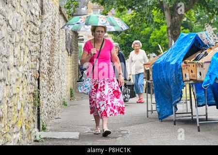 Axminster, Devon, UK.  12th July 2018. UK Weather.  Shoppers in the town of Axminster in Devon with umbrellas up as a light shower passes over on a warm overcast morning.  Picture Credit: Graham Hunt/Alamy Live News Stock Photo