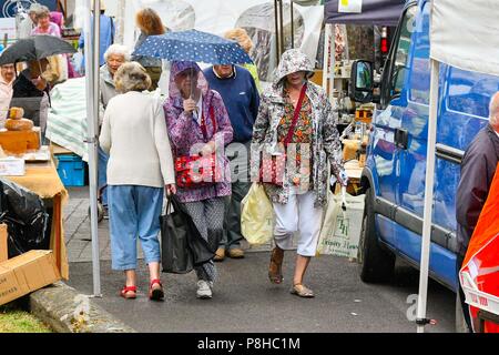 Axminster, Devon, UK.  12th July 2018. UK Weather.  Shoppers in the town of Axminster in Devon with umbrellas up as a light shower passes over on a warm overcast morning.  Picture Credit: Graham Hunt/Alamy Live News Stock Photo