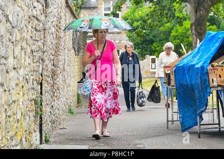 Axminster, Devon, UK.  12th July 2018. UK Weather.  Shoppers in the town of Axminster in Devon with umbrellas up as a light shower passes over on a warm overcast morning.  Picture Credit: Graham Hunt/Alamy Live News Stock Photo