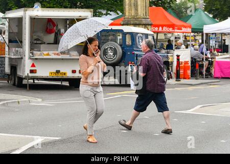 Axminster, Devon, UK.  12th July 2018. UK Weather.  Shoppers in the town of Axminster in Devon with umbrellas up as a light shower passes over on a warm overcast morning.  Picture Credit: Graham Hunt/Alamy Live News Stock Photo