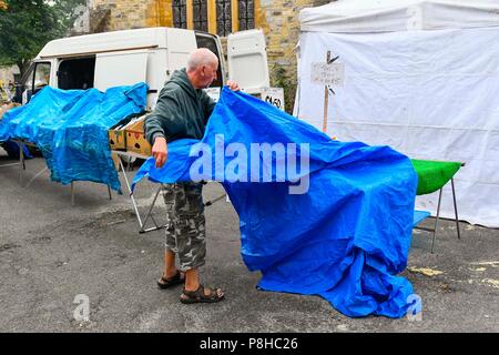 Axminster, Devon, UK.  12th July 2018. UK Weather.  A market stall holder covers his goods with a tarpaulin in the town of Axminster in Devon as a light shower passes over on a warm overcast morning.  Picture Credit: Graham Hunt/Alamy Live News Stock Photo