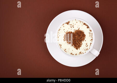 Close up white cup full of latte cappuccino coffee with heart shaped chocolate on milk froth at porcelain saucer over brown paper background, close up Stock Photo
