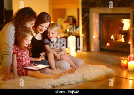 Happy young family using a tablet pc at home by a fireplace in warm and cozy living room on winter day Stock Photo