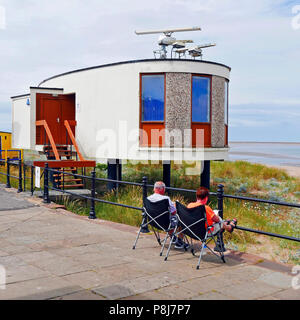 Middle aged couple sat in identical folding chairs reading beneath the Blackpool and Fylde College radar training station on Fleetwood seafront, Stock Photo