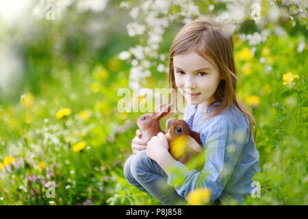 Adorable little girl in blooming cherry garden holding two easter bunnies on beautiful spring day Stock Photo