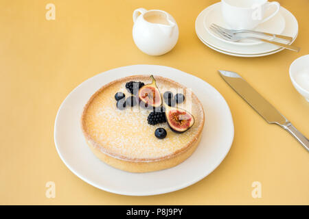 Lemon tart decorated with fresh figs, blueberries and blackberries on a table with plates, cake forks, cup, milk jar and sugar bowl in the background. Stock Photo