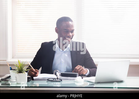 Happy African Businessman Looking At Laptop Calculating Invoice In The Office Stock Photo