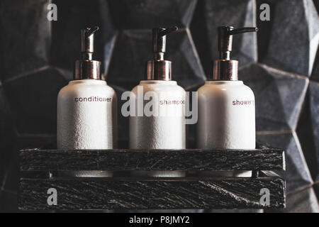 Three white metal bottles of shampoo, conditioner, shower gel on wooden shelf  in modern hotel bathroom with textured black wall Stock Photo
