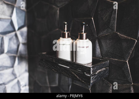 Two white metal bottles of conditioner, shower gel on wooden shelf  in modern hotel bathroom with textured black wall Stock Photo